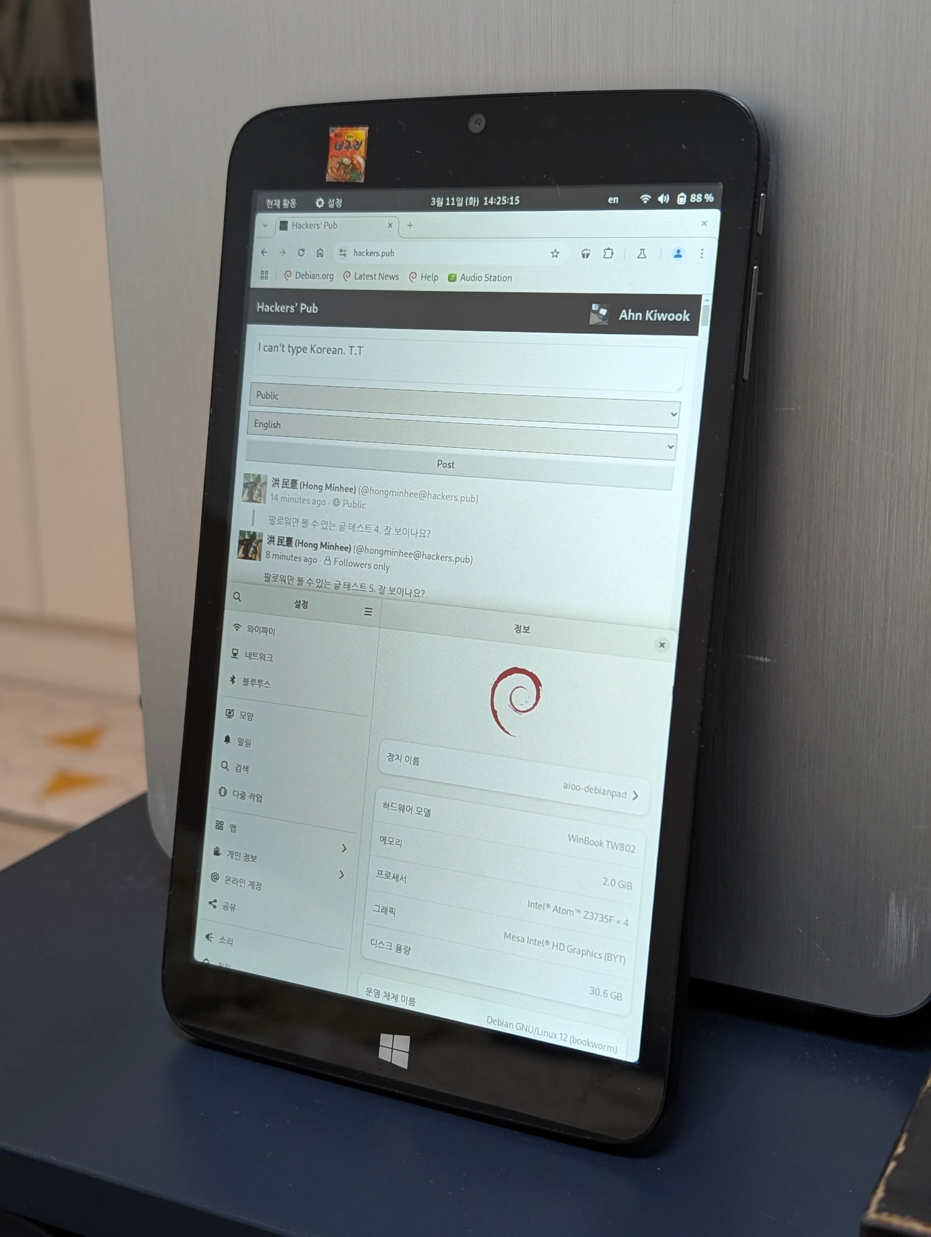 이 이미지는 어두운 표면 위에 세워진 태블릿 기기를 보여주고 있으며, 은색의 데스크탑 앞에 기대어져 놓여 있습니다. 이 기기는 하단에 Windows 로고가 있고 여러 화면을 표시하고 있습니다.
화면 상단에는 "hackers.pub" URL과 탐색 요소가 표시되어 있습니다. 메인 화면에는 "Hackers' Pub"이라는 웹페이지가 표시되어 있으며, 한국어 텍스트와 영어 요소가 포함되어 있습니다. 대화 영역에는 "I can't type Korean. T.T"라는 메시지가 보입니다. 
화면 하단에는 "Winbook TW802", "2.0 GiB" 메모리, "Intel Atom Z3735F", "Mesa Intel HD Graphics", "30.6 GB" 디스크 용량과 같은 기기에 대한 시스템 정보가 표시되어 있습니다. 운영 체제는 "Debian GNU/Linux 12 (bookworm)"인 것으로 보입니다.
기기의 상태 표시줄에는 날짜와 시간(3월 11일 14:25:15), 배터리 수준(88%) 및 연결 아이콘이 표시되어 있습니다. 또한 기기 상단에 작은 너구리 라면 스티커가 보입니다.