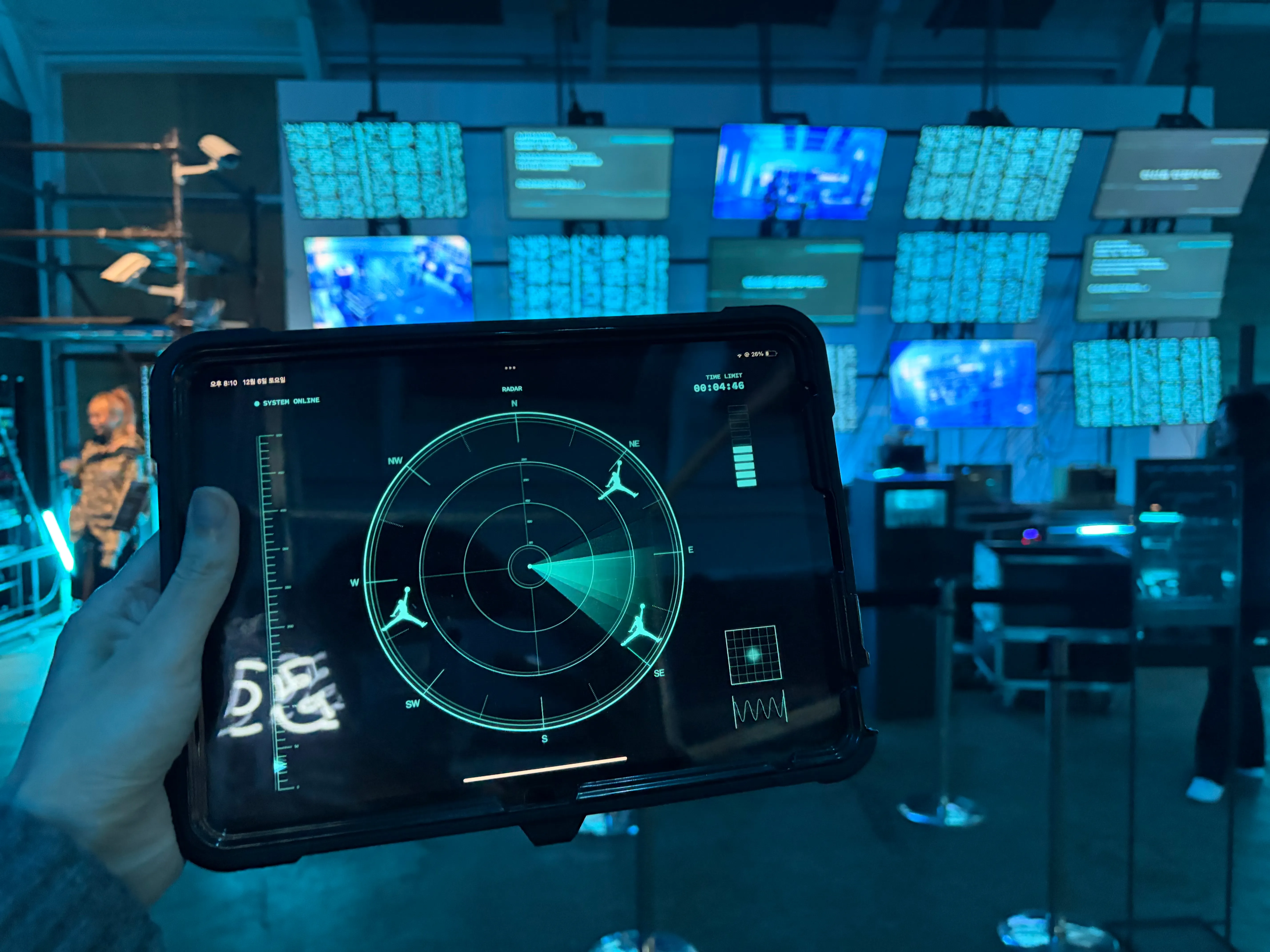 파란 조명의 이벤트 행사장을 배경으로 손에 아이패드가 들려있다. 아이패드 화면에는 레이더망과 조던 로고가 보인다.